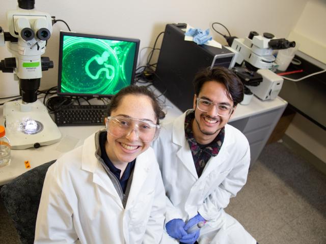 Two students in lab coats and goggles smile in a research lab with microscopes and a glowing image on a computer screen.