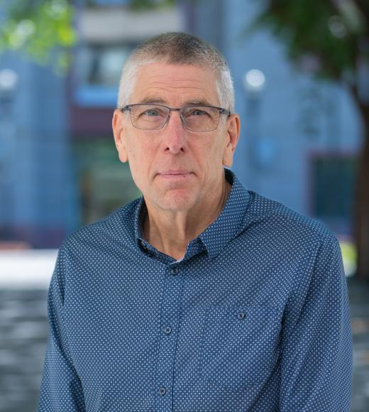 Outdoor headshot of older man with gray hair, glasses, blue shirt, neutral expression