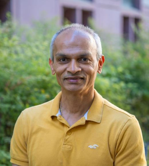 Person smiling outdoors in a mustard-yellow polo shirt, short gray hair, greenery and building background.
