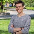 A person with short hair and a gray long-sleeve shirt stands smiling with arms crossed on a sunny, tree-lined lawn at UC Davis.