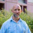 Person smiling outdoors wearing a light blue embroidered shirt, bald with beard, greenery behind.