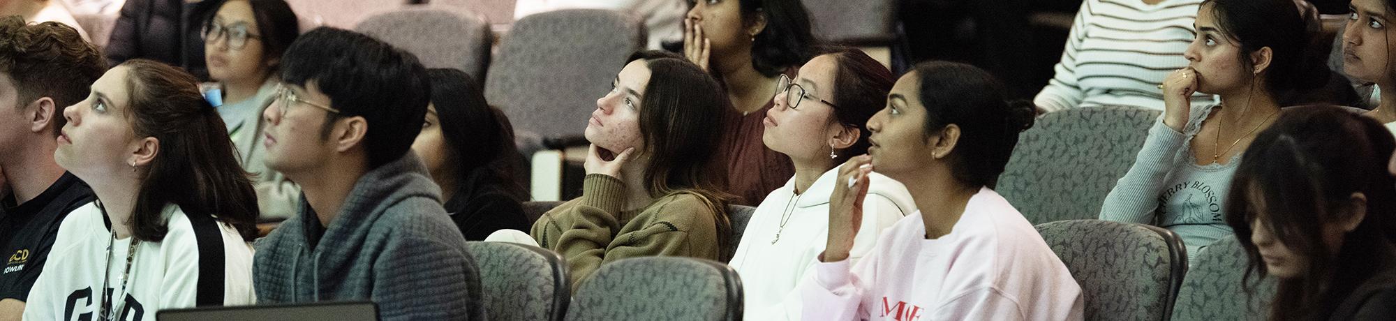 A group of college students sit in an auditorium, all facing toward the front with attentive expressions. Most students have their chins resting on their hands or fingers to their faces, suggesting deep focus. The lighting is soft and academic, and the seating consists of padded gray chairs. The students represent a diverse group, and several are wearing sweatshirts or casual clothing. The atmosphere suggests they are watching a presentation or lecture.