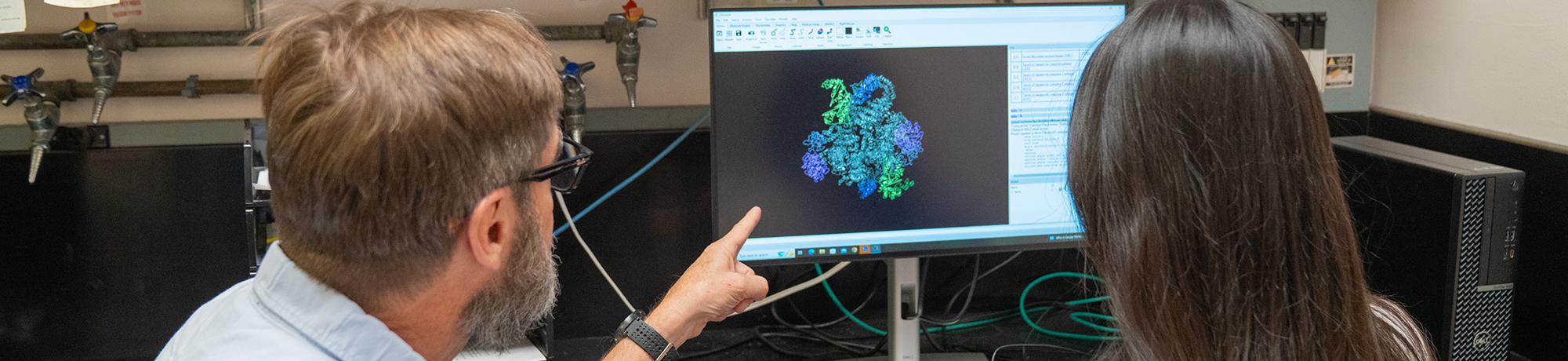 A man and a woman sit at a computer in a lab, viewing a colorful 3D molecular structure on the monitor. The man is pointing at the screen while explaining something. Lab equipment and tubing are visible in the background.