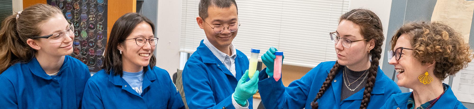 Five students and one instructor wearing blue lab coats and safety glasses gather around in a lab, smiling and observing two colorful sample tubes—one yellow and one pink—held up by one of the students. Lab equipment and a chart are visible in the background.