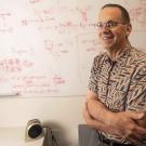 Man smiling in front of whiteboard with equations, wearing glasses.